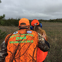 Two people wearing orange practice firearm safety in a woodland prairie.
