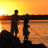 The sun creates a silhouette of two people fishing in a boat on a lake.